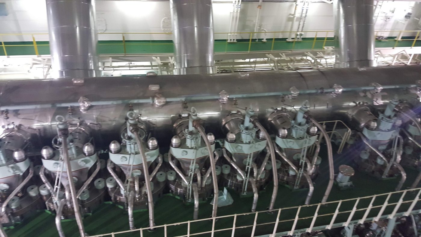Top of a large two-stroke engine: a row of cylinder covers with the fuel injectors fed by lagged high-pressure pipes, and the exhaust receiver running behind